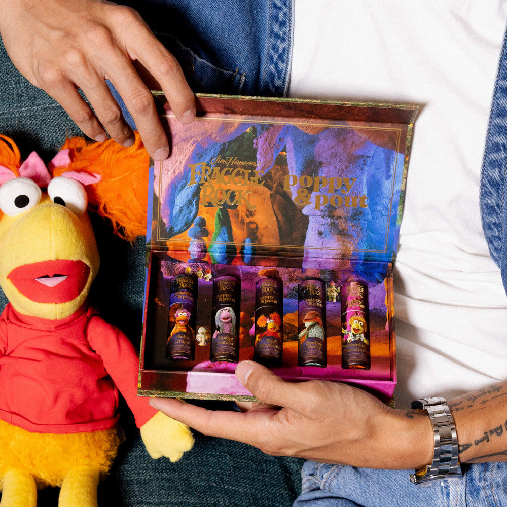 Person holding a Fraggle Rock-themed Poppy & Pout lip balm set with a plush Fraggle character nearby.