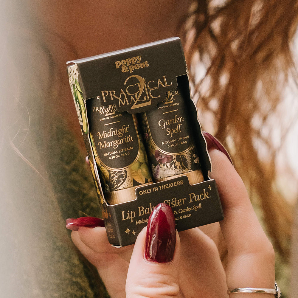 A person holds a boxed lip balm sister pack. The packaging is black with gold lettering and shows two lip balms: Midnight Margarita and Garden Spell.