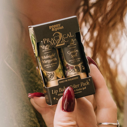 A person holds a boxed lip balm sister pack. The packaging is black with gold lettering and shows two lip balms: Midnight Margarita and Garden Spell.